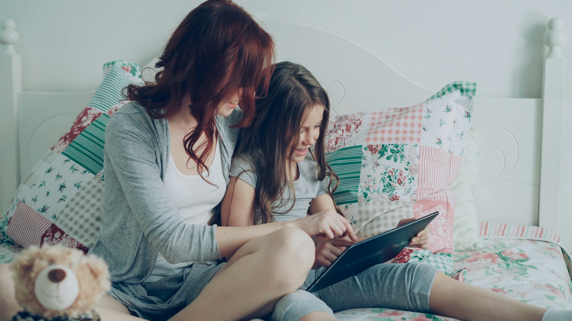 Two girls sitting on a bed with patterned pillows, looking at a tablet together.